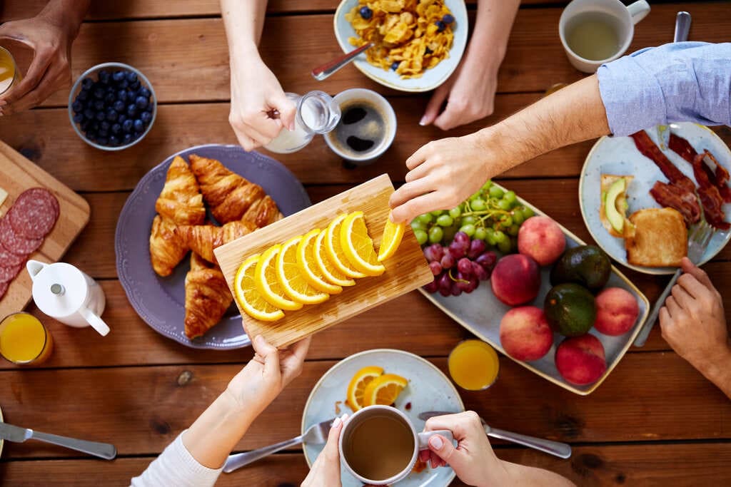 Café da manhã compartilhado com croissants, frutas frescas, cereais, suco de laranja, café e chá sobre uma mesa de madeira.